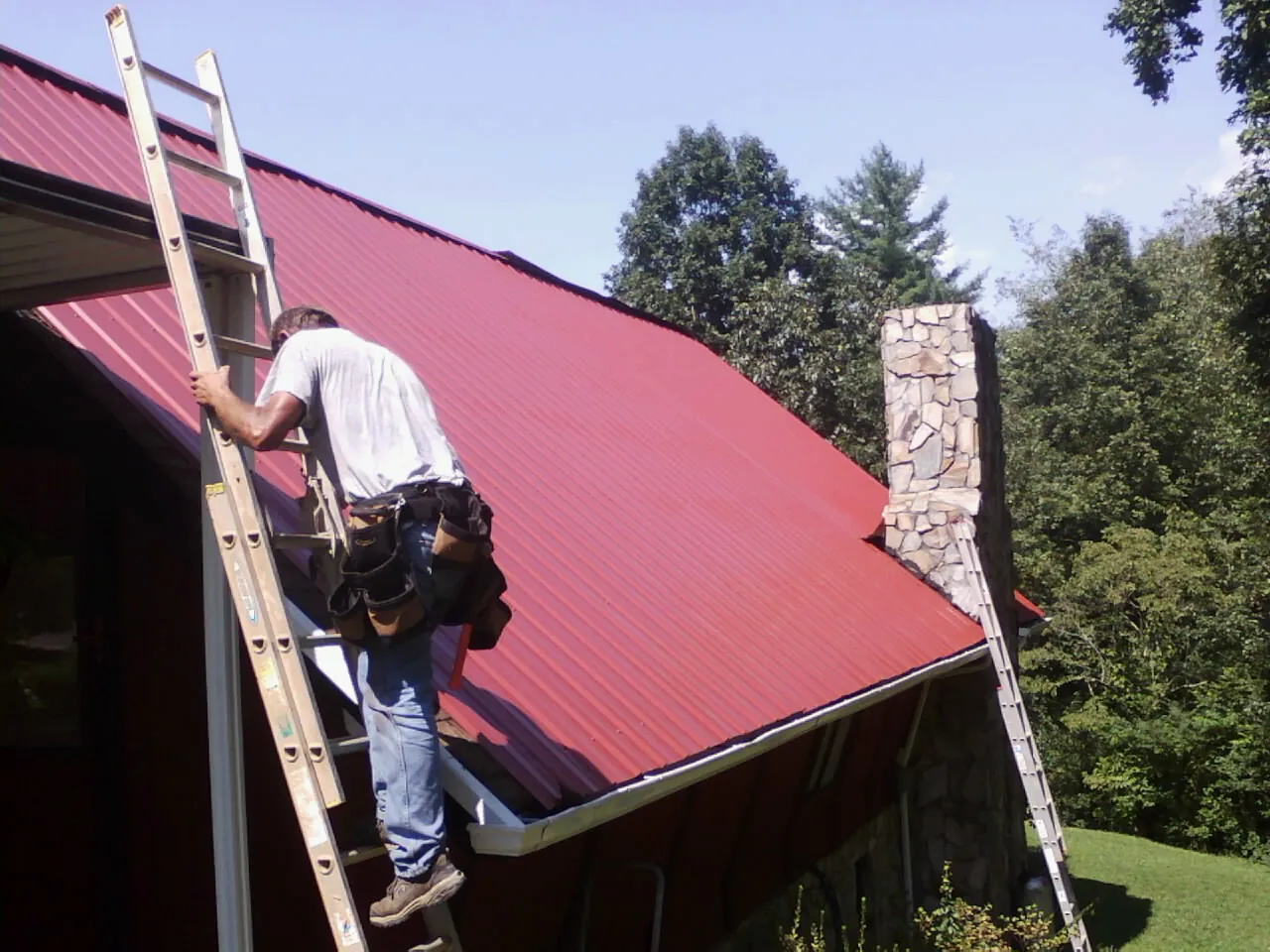 Roofer on a ladder beside a red metal roof and stone chimney — Soffit & Fascia Repair by Roofing Brothers in Richeyville